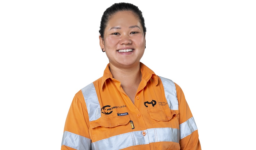 Female civil construction apprentice, wearing orange hi-ves, smiles to the camera.