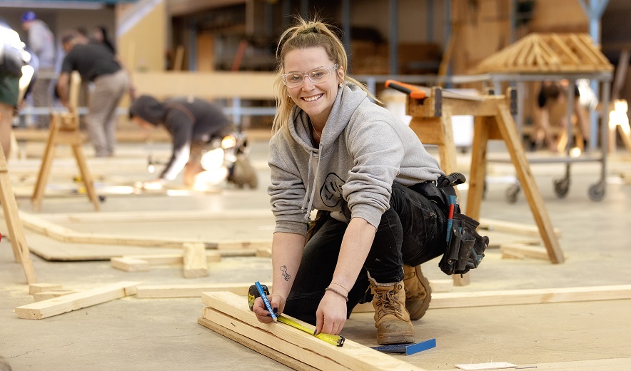 Female apprentice measures a plank of wood in a training workshop.