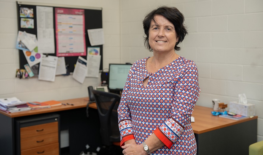 Female teacher stands in office, with desk in background.