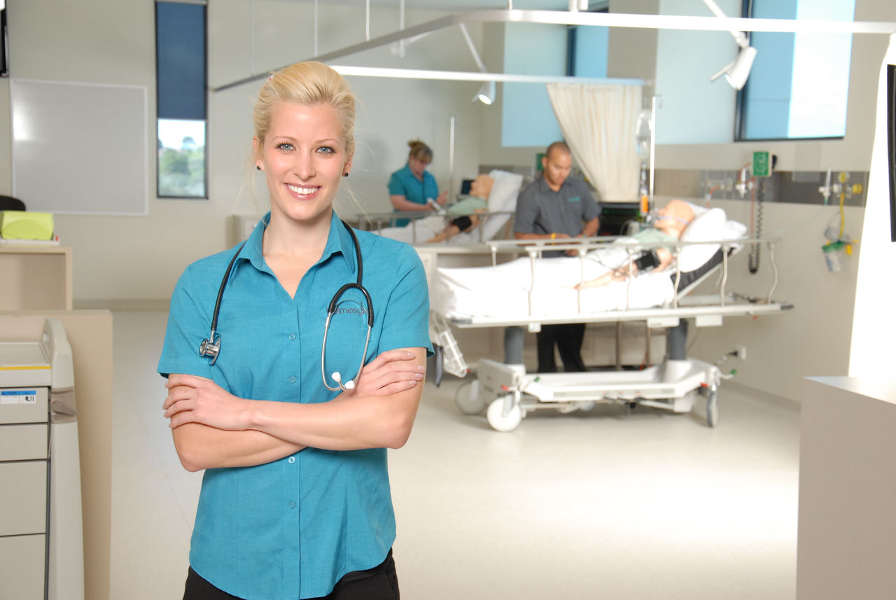 Smiling nursing student stands in training ward, with fellow students in background working on text equipment.