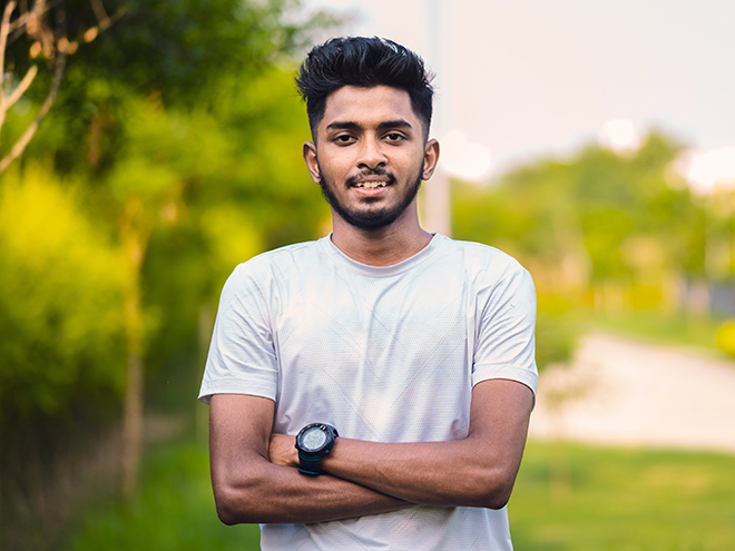 Outdoor Portrait of young Asian/ Indian athlete wearing sports dress and smartwatch crossed his arms & looking at camera in park with greenery in background.