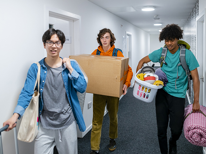 A wide-angle view of a group of three male students who are also friends moving into their new student accommodation. They are chatting with each other and carrying their belongings through the corridor.